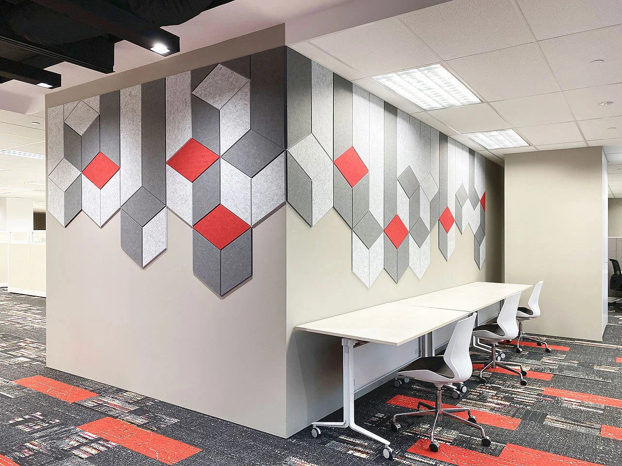 Modern office space with white desks and chairs next to a wall decorated with geometric gray and red PET polyester panels. The carpet has matching gray, black, and red patterns, and the space is brightly lit with overhead lights.