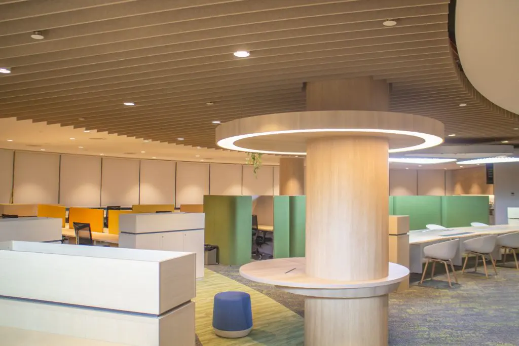 Modern open-plan office with wooden accents, green PET polyester panel partitions, desk chairs, white cabinets, and a circular light fixture around a central column. The space is well-lit and features a contemporary, minimalist design.
