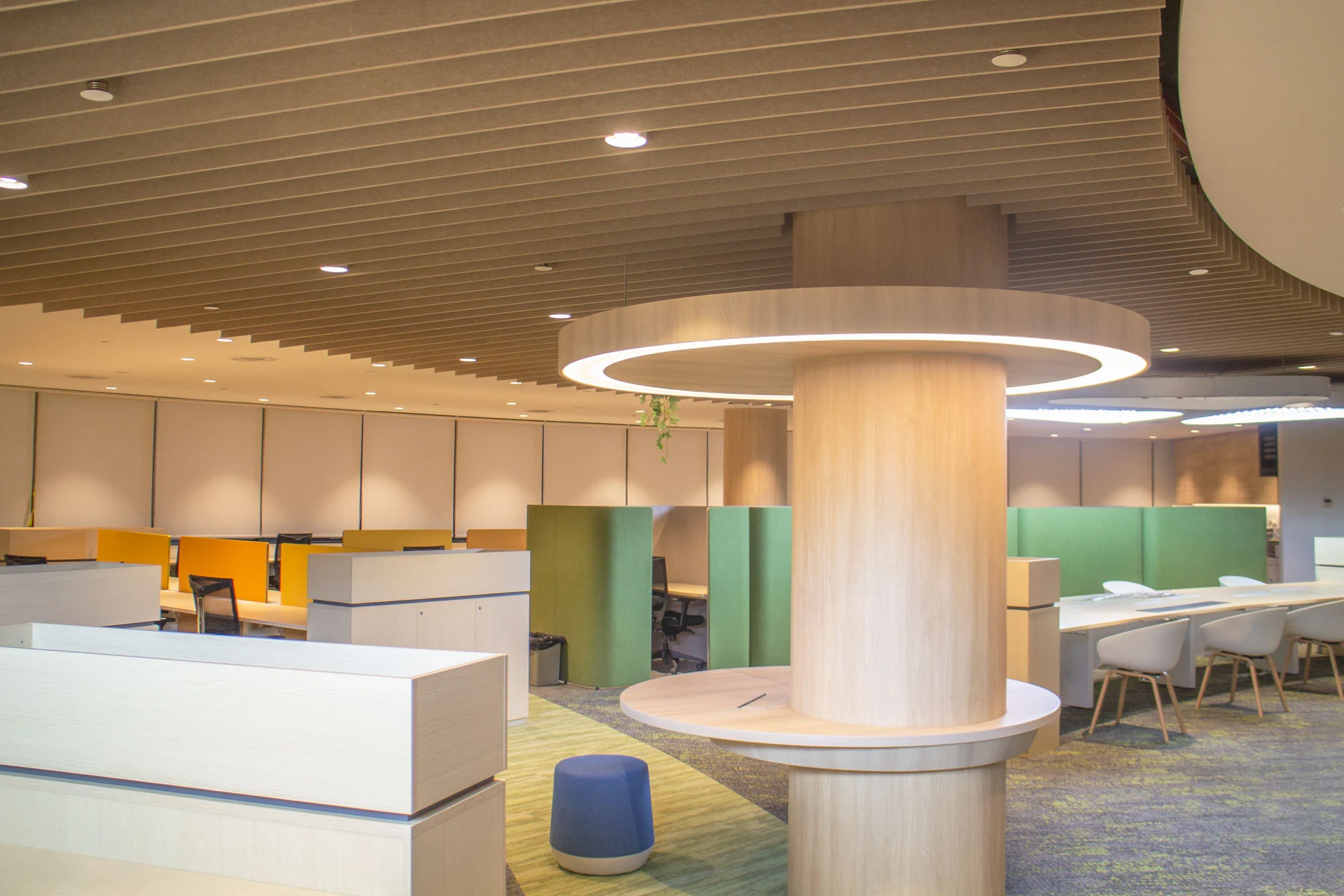 Modern office space with wooden accents, circular overhead lighting, and partitioned workstations in yellow and green using PET polyester panels. A mix of chairs and stools sit on a carpeted floor, creating a bright and organized atmosphere.