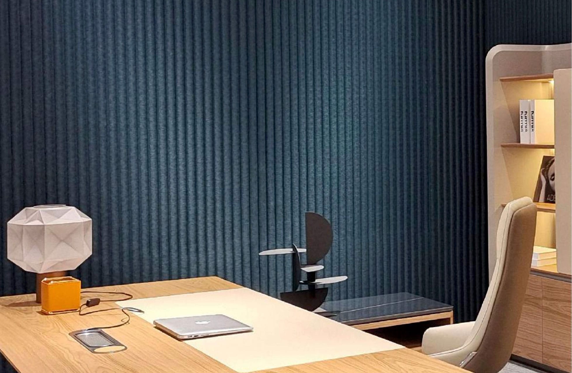 A modern office with a wooden desk, closed laptop, geometric lamp, chair, and bookshelf. Polyx Curved Acoustic Panels accent the textured dark blue wall, creating a neat, well-organized space with enhanced sound control.