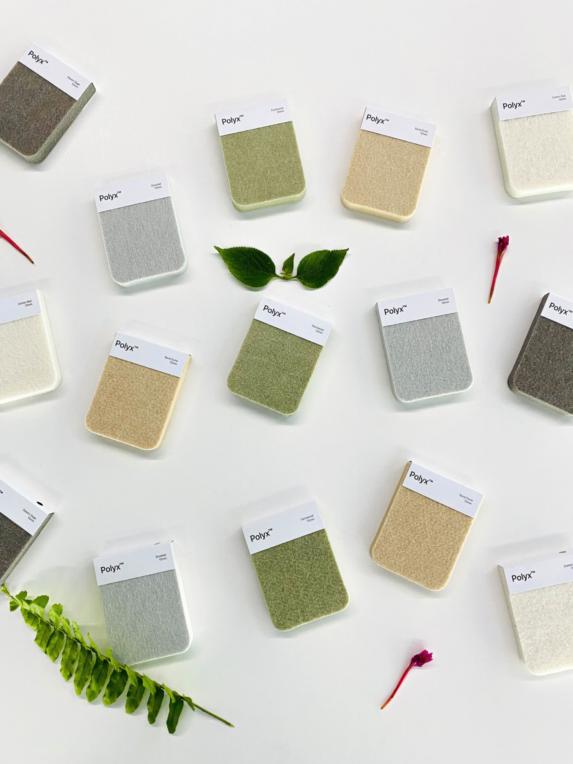 Rectangular fabric swatches in neutral and pastel shades are arranged on a white surface, surrounded by green leaves and petals—ideal for designing acoustically balanced retail spaces. Each swatch features a crisp white label.