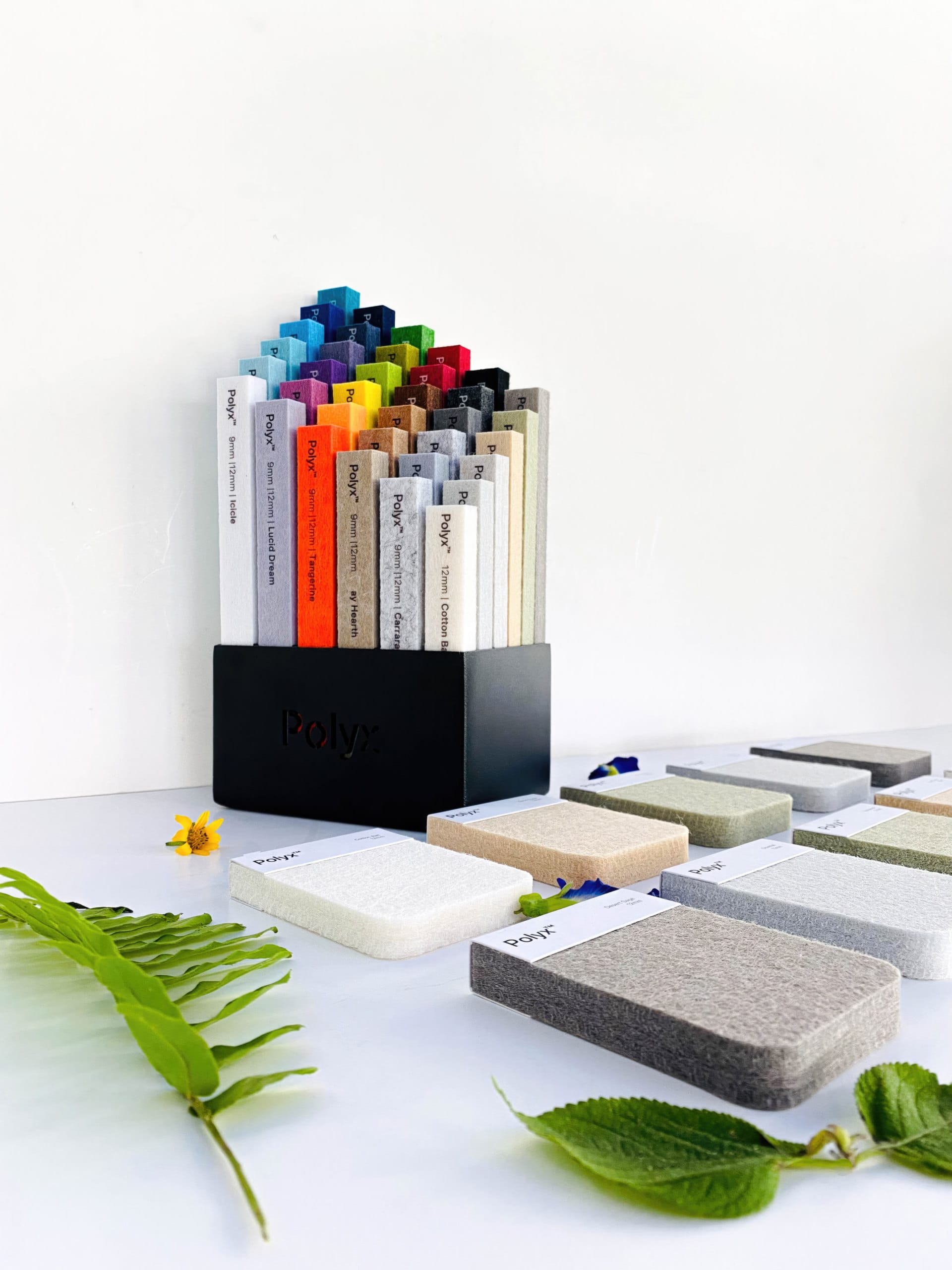 A black holder displays colorful rectangular sample swatches, ideal for designing acoustically balanced retail spaces. More swatches lay flat nearby, while green leaves and a yellow flower rest on the white surface.