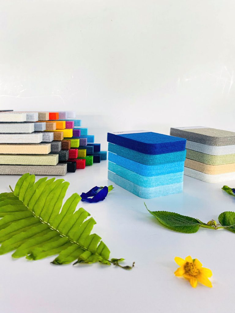 Colorful rectangular sponges are stacked in groups on a white surface, surrounded by a green fern leaf, a blue flower petal, and a yellow flower—perfect for adding a touch of nature to acoustically balanced retail spaces.