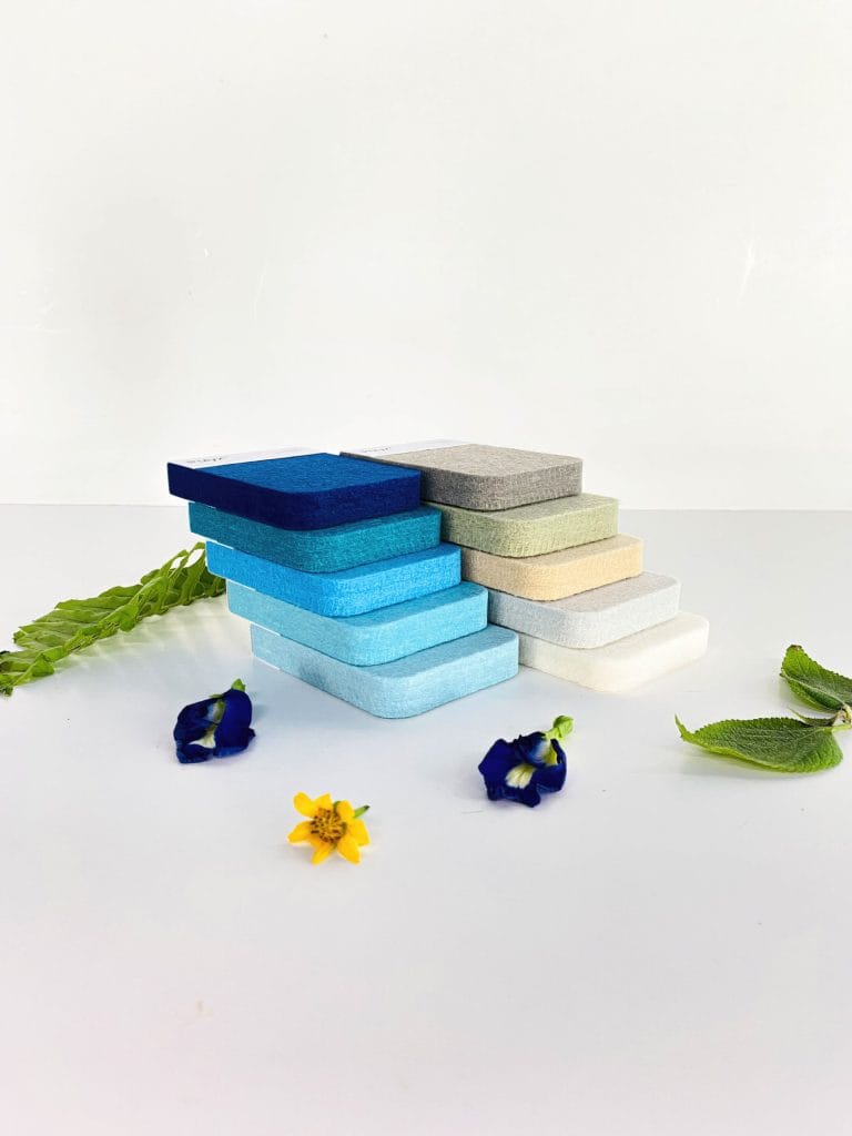 Rectangular soap bars in shades of blue, gray, green, and beige are stacked on a white surface in a stepped display, evoking the calm design of acoustically balanced retail spaces. Green leaves and small yellow and purple flowers surround the soaps.
