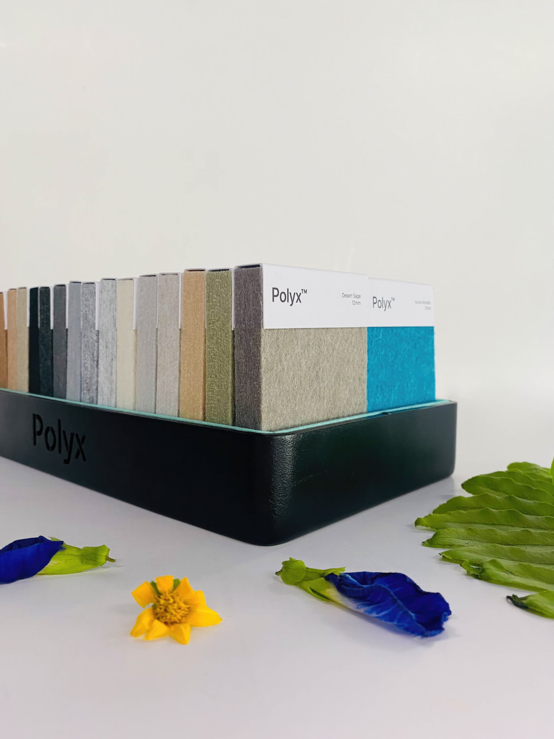 A display of Polyx material samples in various colors, organized upright in a black holder, ideal for acoustically balanced retail spaces, with a yellow flower, blue petals, and green leaves arranged on a white surface nearby.