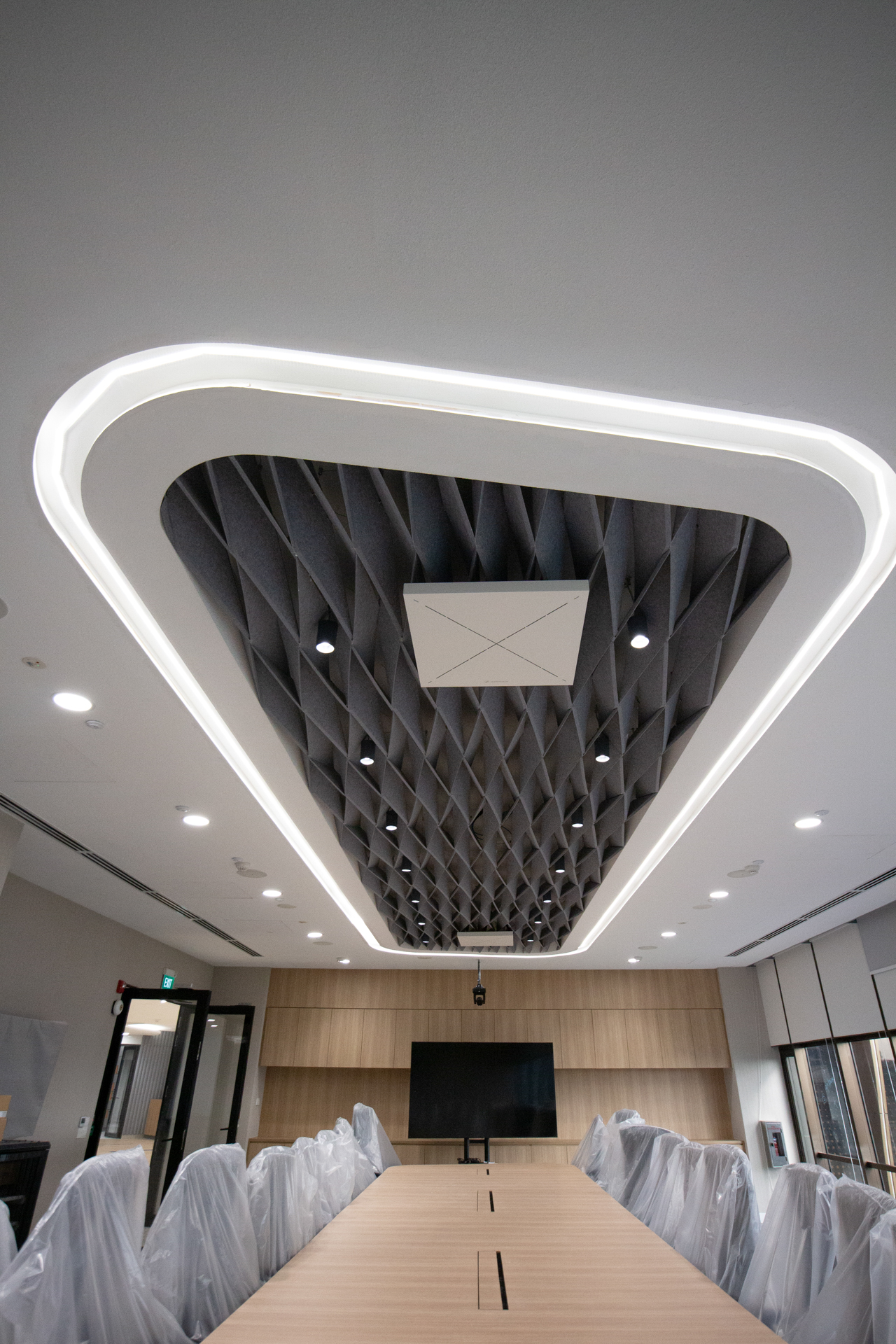 A modern conference room with a long wooden table, chairs covered in plastic, a large wall-mounted screen, and a ceiling with geometric acoustic panels and recessed lighting.