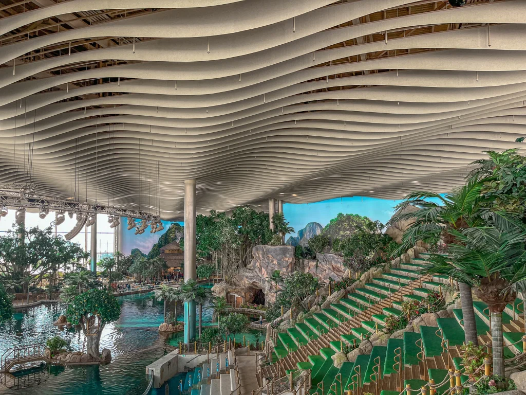 Indoor water park with a wavy, modern ceiling, artificial tropical trees, rock structures, a large pool, and green seating steps along one side. The atmosphere is bright and lush.