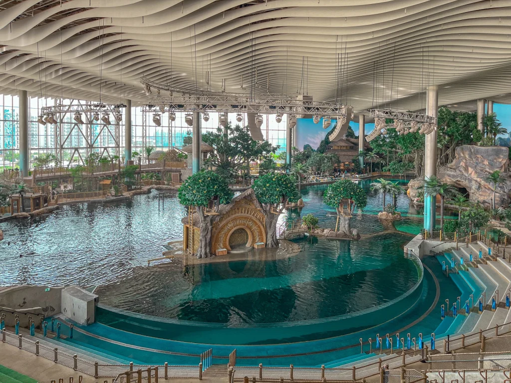 A spacious indoor water park with a large pool, decorative structures resembling ancient ruins, artificial trees, and tropical plants, all under a high, wavy ceiling with large windows letting in natural light.