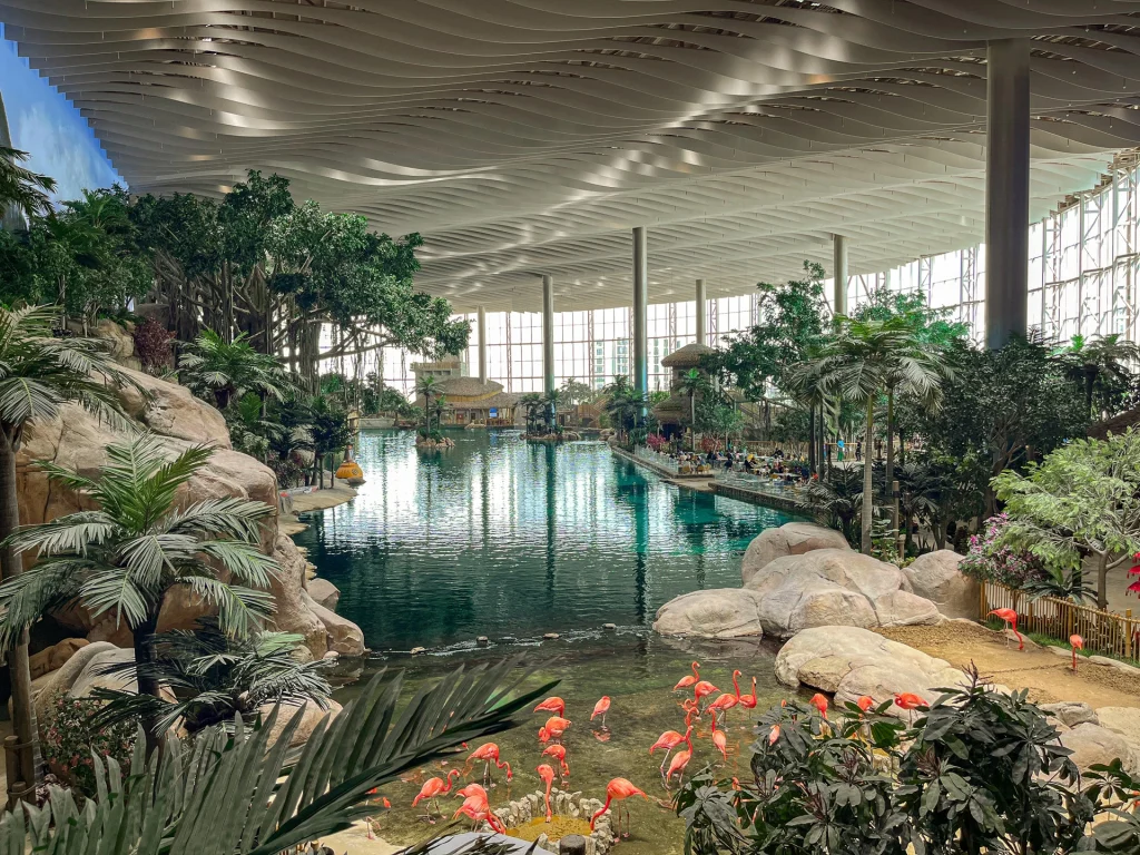A large indoor tropical paradise with lush green plants, a lagoon, flamingos in the foreground, and sunlight streaming through tall glass windows and a wavy-patterned ceiling.