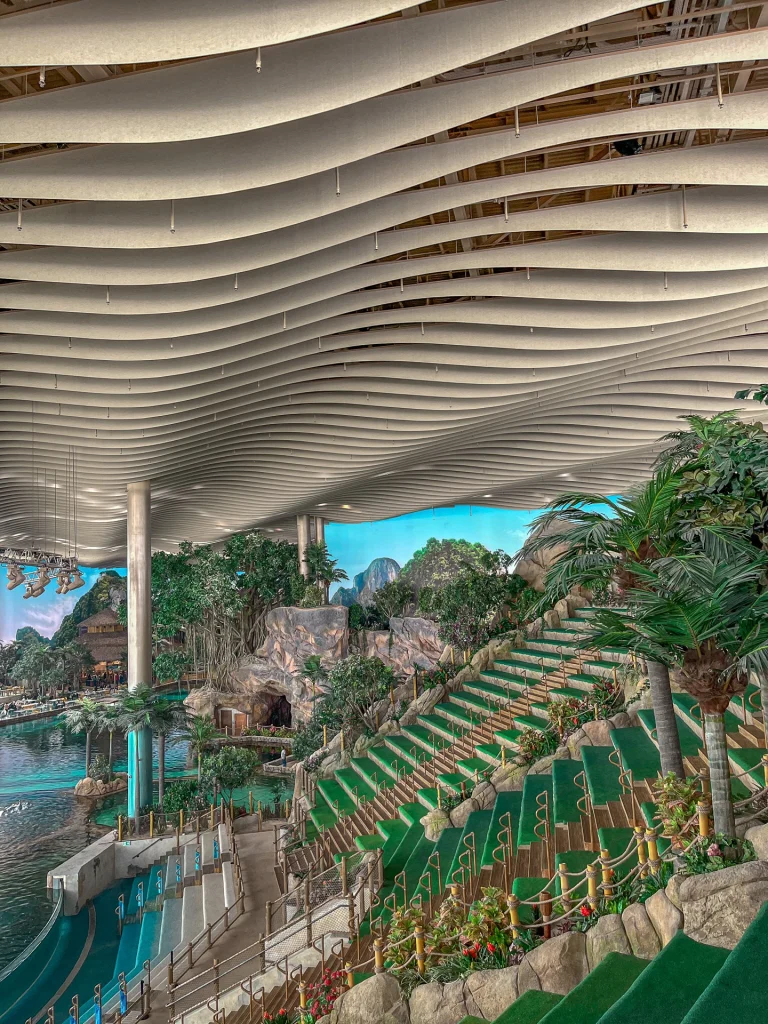 Indoor water park with tiered green seating, lush artificial plants, a large pool, and wavy ceiling panels. Tropical decorations and rock features create a resort-like, immersive atmosphere.
