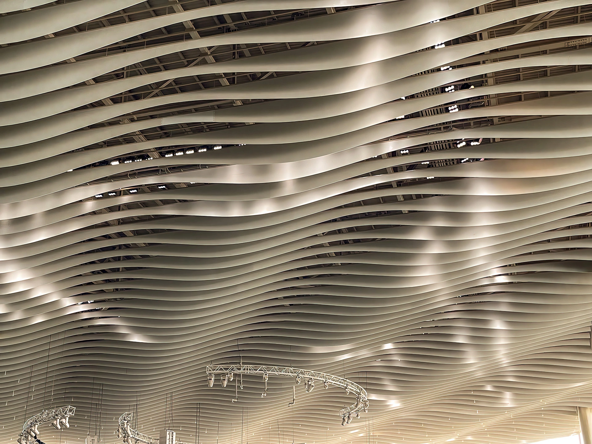 Curved metallic panels form a wavy ceiling with lighting reflecting off the surface. Below, there are circular truss structures and visible beams, creating an architectural and modern design.