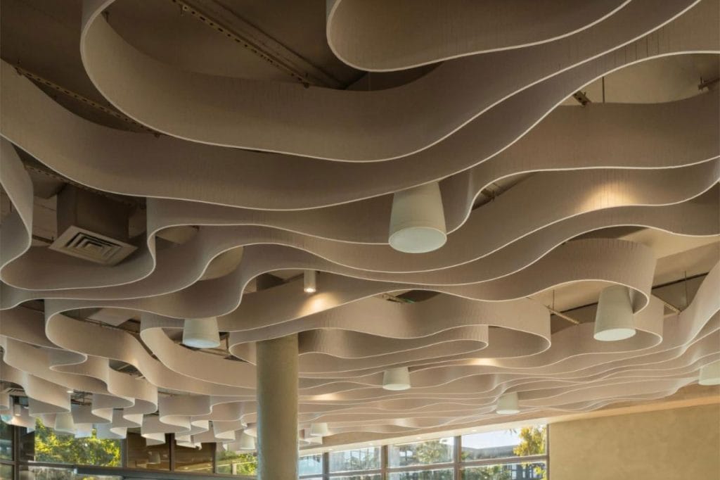 Curved, wave-like ceiling panels with integrated cylindrical white lights hang in a modern room with large windows and a pillar. Natural light filters in, highlighting the unique architectural design.