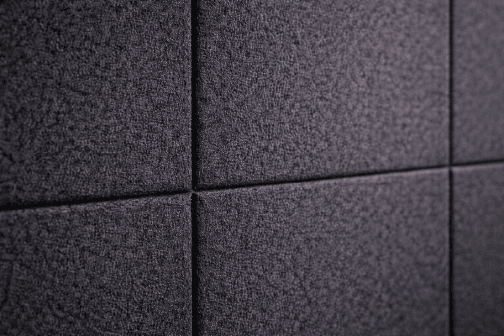 Close-up of a wall made of dark gray, textured acoustic panels arranged in a grid pattern, with visible seams between the rectangular panels.