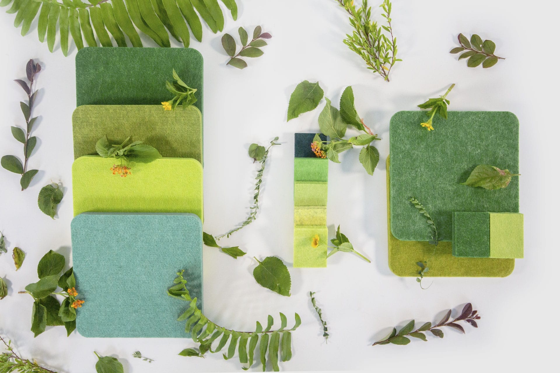 Green and yellow fabric swatches arranged with rounded corners, surrounded by various green leaves and small yellow flowers on a white background. The scene highlights nature-inspired colors and textures.