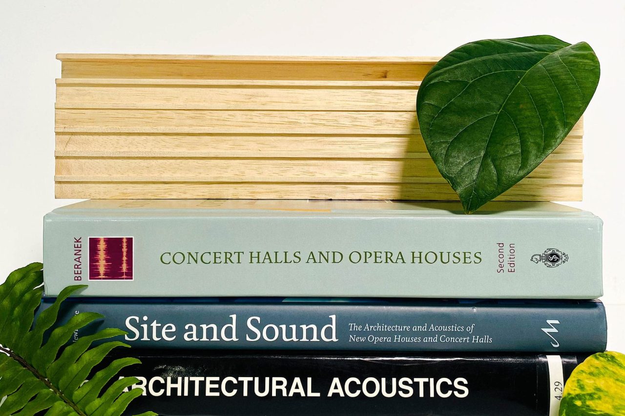 Three books—Concert Halls and Opera Houses, Site and Sound, and Architectural Acoustics—are stacked beside green leaves, with a wooden object behind them, reflecting the inspiration behind Our Other Acoustical Products.