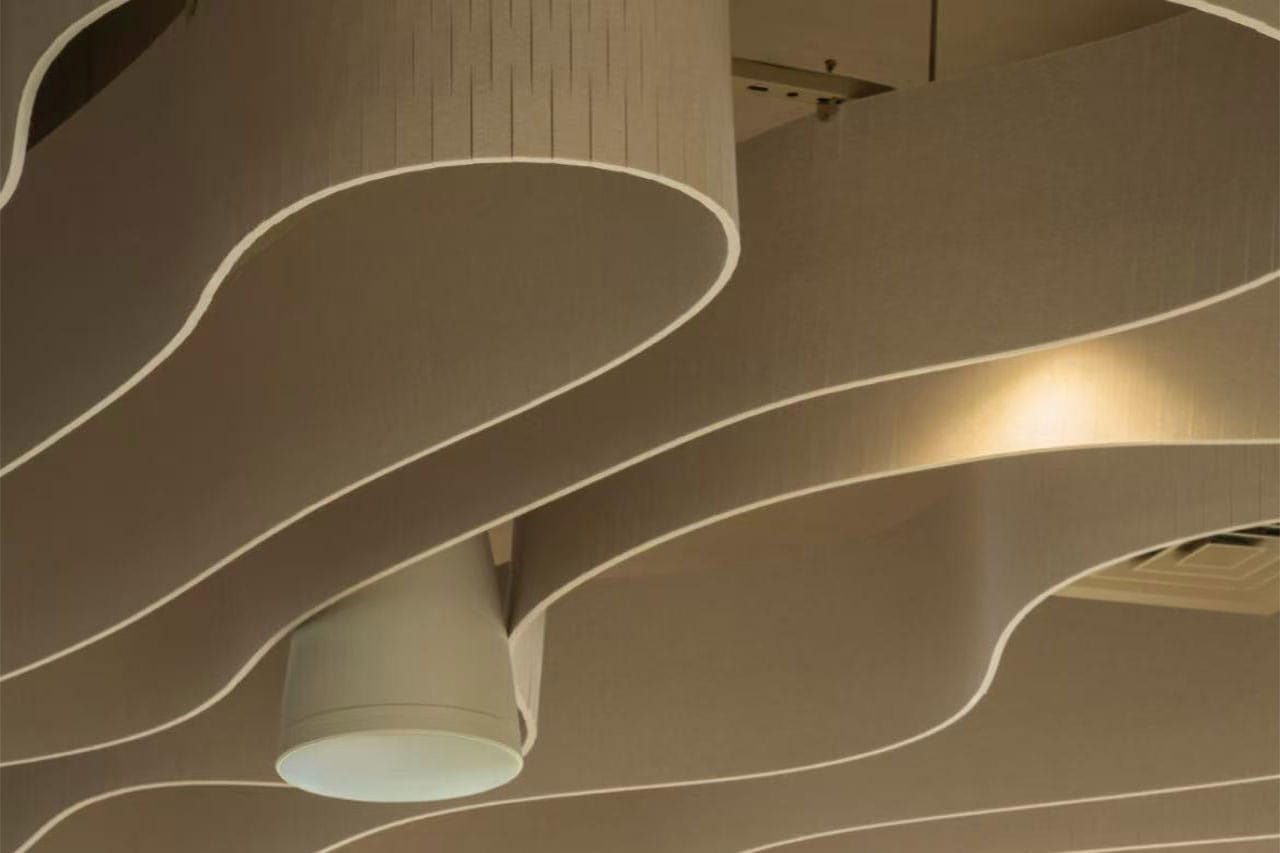 Curved, wave-like architectural ceiling panels with a cylindrical white light fixture and a warm spotlight illuminating part of the surface. The design features soft, flowing lines and neutral beige colors.