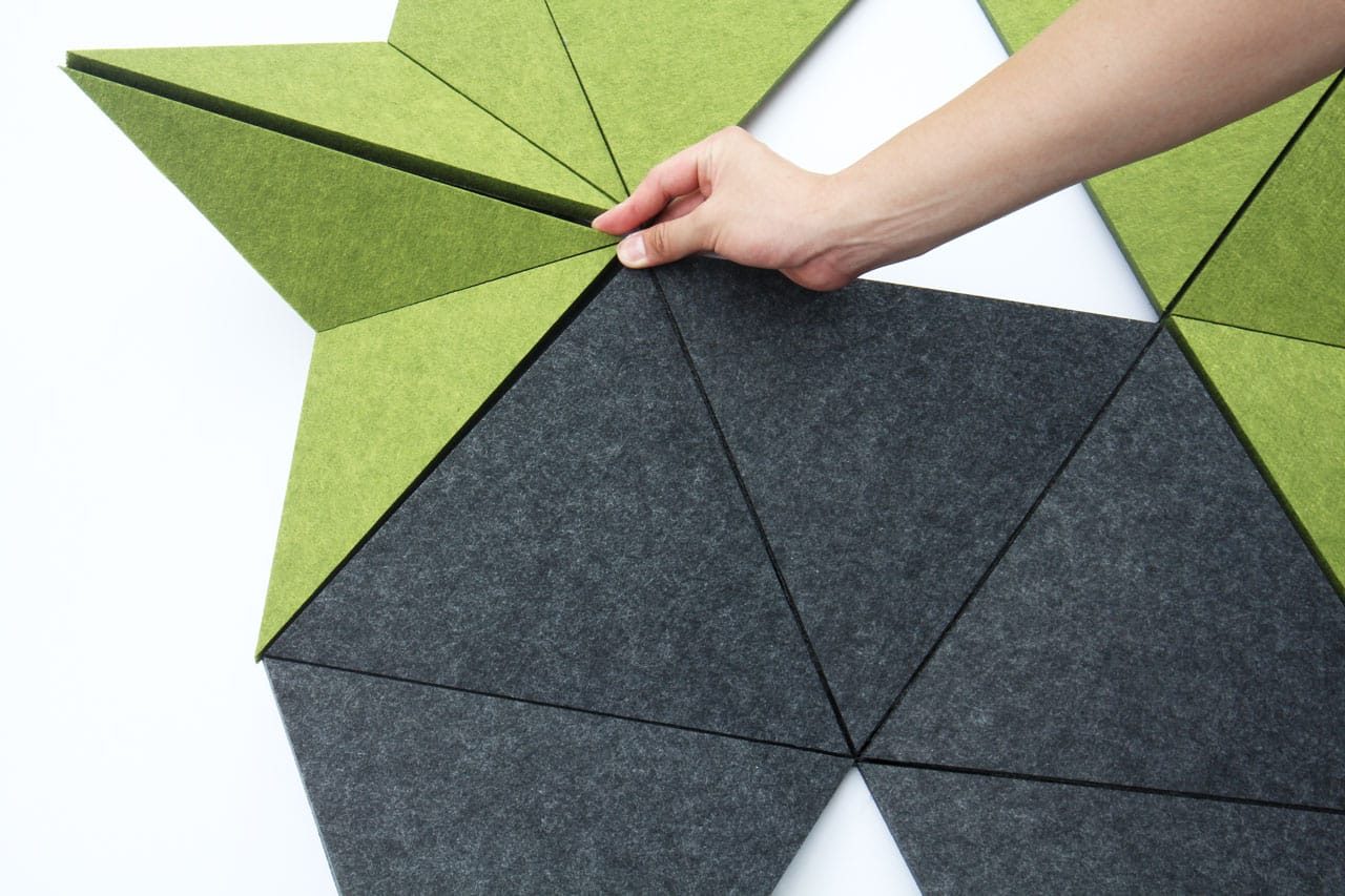 A hand arranges green and dark gray triangular polyester acoustic panels offerings on a white surface, forming a geometric pattern.
