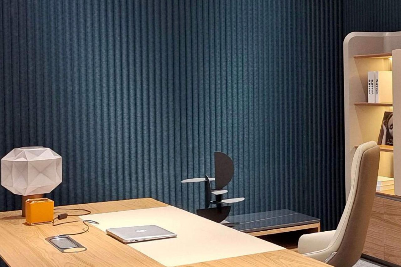 A modern office with a wooden desk, closed laptop, geometric lamp, chair, and bookshelf. Polyx Curved Acoustic Panels accent the textured dark blue wall, creating a neat, well-organized space with enhanced sound control.